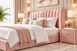 A blush pink velvet bed with a tall, channel-tufted headboard, styled with white bedding, patterned accent pillows, and a matching pink nightstand against a black-and-white botanical wallpaper.