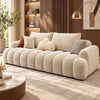 A modern beige tufted sofa with chunky rounded arms and back cushions, styled in a warm contemporary living room with wood paneling, a gold floor lamp, and neutral throw pillows.
