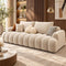 A modern beige tufted sofa with chunky rounded arms and back cushions, styled in a warm contemporary living room with wood paneling, a gold floor lamp, and neutral throw pillows.