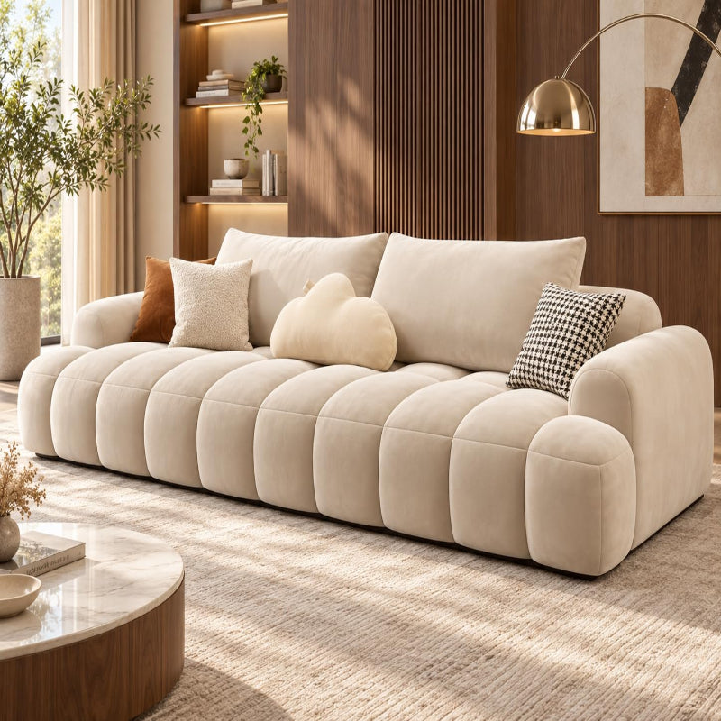 A modern beige tufted sofa with chunky rounded arms and back cushions, styled in a warm contemporary living room with wood paneling, a gold floor lamp, and neutral throw pillows.